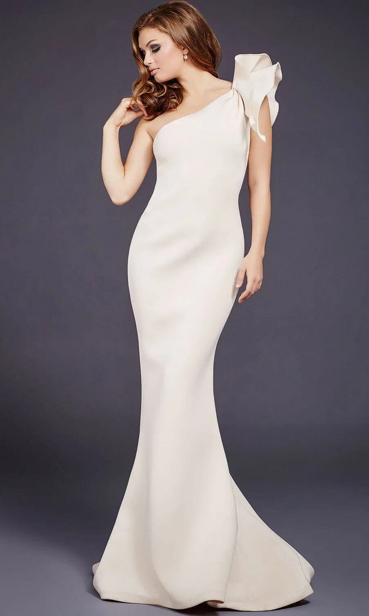 A woman poses against a plain dark background wearing the Jovani 32602 One-Shoulder Mermaid Prom Gown, a fitted, floor-length off-white dress featuring a single shoulder and a statement bow accent.